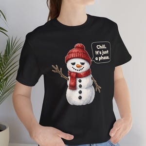 Pode incluir: Uma t-shirt preta com o gr&aacute;fico de um boneco de neve usando um gorro e cachecol de malha vermelhos. O boneco de neve tem bra&ccedil;os de galho, nariz de cenoura e um bal&atilde;o de fala com o texto: Chill. It's just a phase.