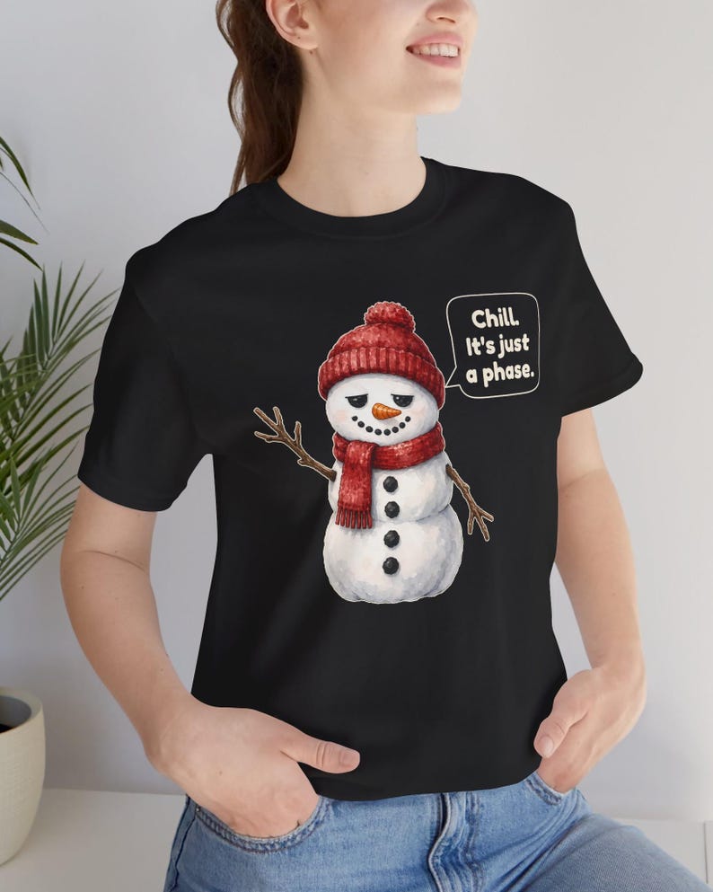 Pode incluir: Uma t-shirt preta com o gr&aacute;fico de um boneco de neve usando um gorro e cachecol de malha vermelhos. O boneco de neve tem bra&ccedil;os de galho, nariz de cenoura e um bal&atilde;o de fala com o texto: Chill. It's just a phase.