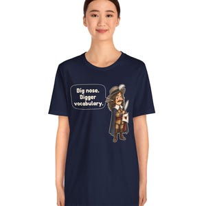 May include: A navy blue short-sleeve t-shirt featuring a graphic of a historical figure with a large nose, wearing a hat and cape, holding a quill and letter. A speech bubble contains the text: Big nose. Bigger vocabulary.