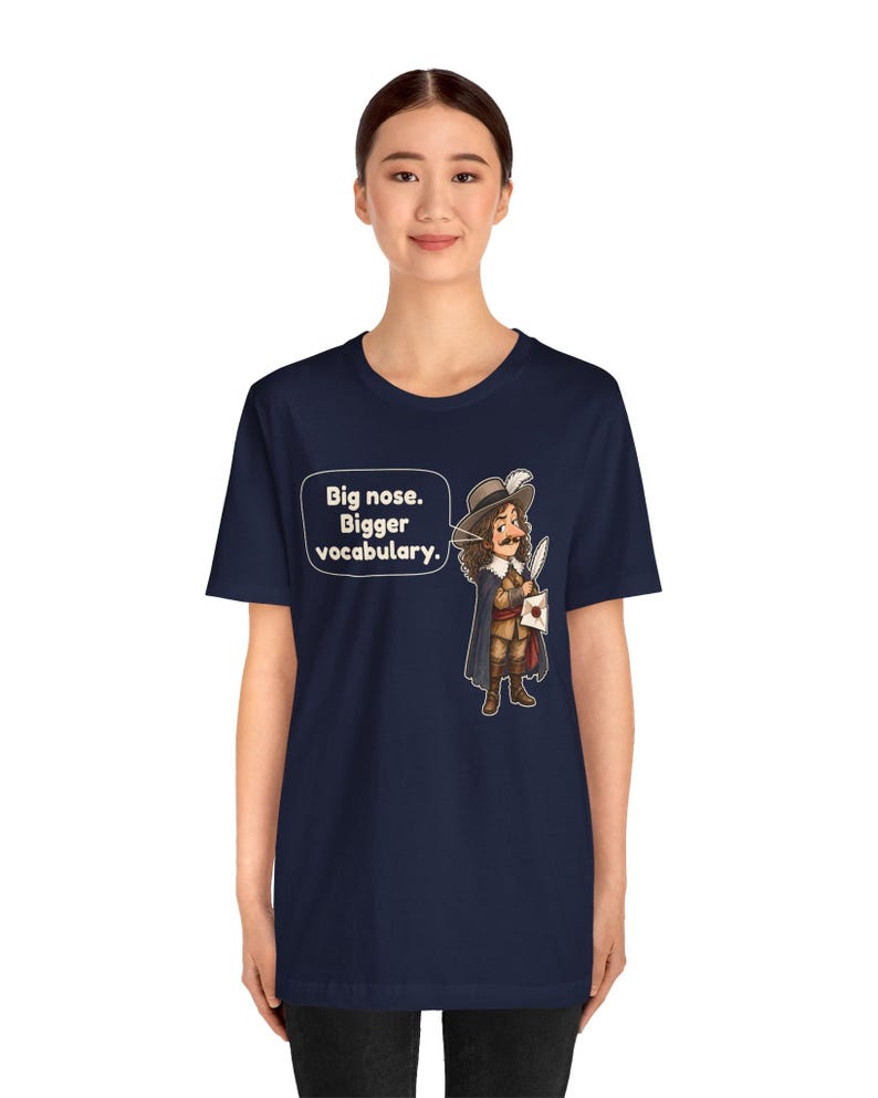 May include: A navy blue short-sleeve t-shirt featuring a graphic of a historical figure with a large nose, wearing a hat and cape, holding a quill and letter. A speech bubble contains the text: Big nose. Bigger vocabulary.