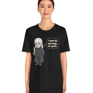 May include: A black short-sleeve t-shirt featuring a graphic of a pale, long-haired character in a dark coat holding a lit candle. A speech bubble next to the character contains the text: I don't do mornings. Or garlic.
