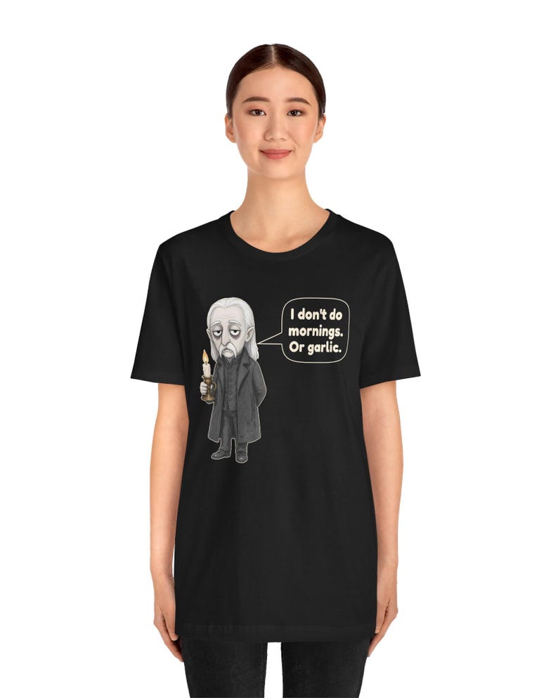 May include: A black short-sleeve t-shirt featuring a graphic of a pale, long-haired character in a dark coat holding a lit candle. A speech bubble next to the character contains the text: I don't do mornings. Or garlic.