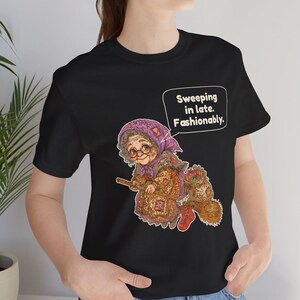 Puede incluir: Una camiseta negra de manga corta con el gr&aacute;fico de una persona mayor con pa&ntilde;uelo morado y gafas montada en una escoba. El dise&ntilde;o incluye un bocadillo con el texto "Sweeping in late. Fashionably." y un calcet&iacute;n navide&ntilde;o rojo.