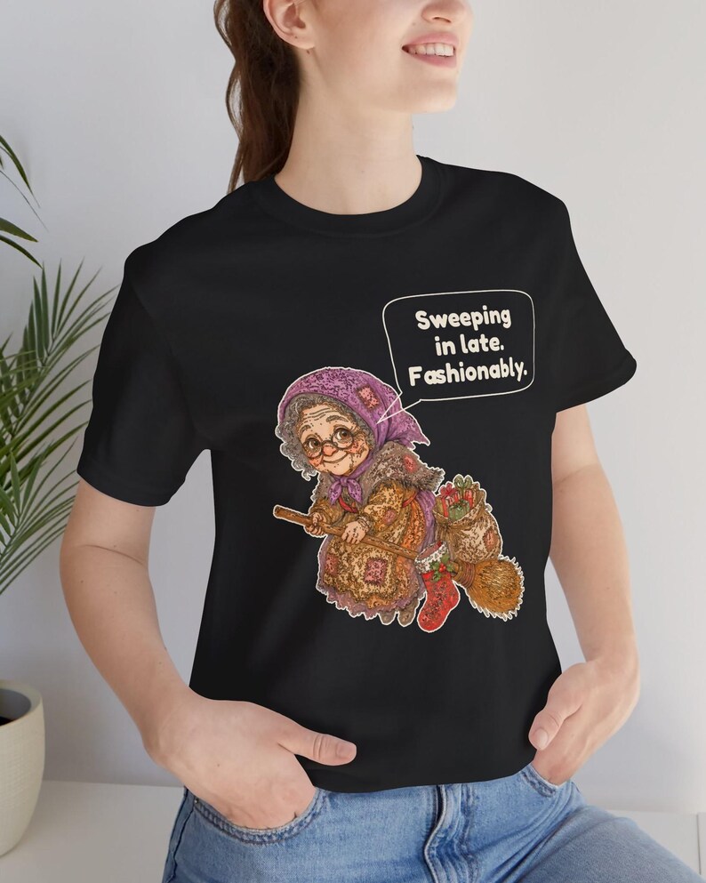 Puede incluir: Una camiseta negra de manga corta con el gr&aacute;fico de una persona mayor con pa&ntilde;uelo morado y gafas montada en una escoba. El dise&ntilde;o incluye un bocadillo con el texto "Sweeping in late. Fashionably." y un calcet&iacute;n navide&ntilde;o rojo.