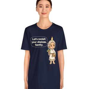 May include: A navy blue t-shirt featuring a graphic of a small, light-haired figure with a candle flame on their head, holding a sprig of holly and a small basket. A speech bubble next to the figure contains the text: Let's revisit your choices. Gently.