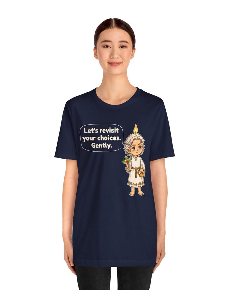 May include: A navy blue t-shirt featuring a graphic of a small, light-haired figure with a candle flame on their head, holding a sprig of holly and a small basket. A speech bubble next to the figure contains the text: Let's revisit your choices. Gently.