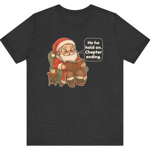 Op de afbeelding: Een donkergrijs gem&ecirc;leerd t-shirt met een afbeelding van de Kerstman die in een groene fauteuil zit en een boek leest, met een klein bijzettafeltje en een mok. Een tekstballon bevat de tekst: Ho ho hold on. Chapter ending.