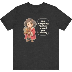 May include: A charcoal gray t-shirt featuring a cartoon illustration of a person with curly brown hair wearing a red coat, holding a golden armillary sphere. A speech bubble next to the figure contains the text: Not everything revolves around you. Literally.
