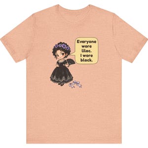May include: A peach-colored t-shirt featuring a graphic of a character in a black dress with a lilac flower crown and fan. A speech bubble contains the text: Everyone wore lilac. I wore black. A small lilac ribbon lies on the ground below.