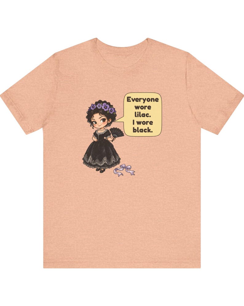 May include: A peach-colored t-shirt featuring a graphic of a character in a black dress with a lilac flower crown and fan. A speech bubble contains the text: Everyone wore lilac. I wore black. A small lilac ribbon lies on the ground below.