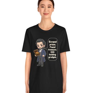 May include: A black t-shirt featuring a cartoon illustration of a bearded man in a blue coat holding a treasure chest. A speech bubble next to the character contains the text: Escaped prison. Found treasure. Still holding grudges.