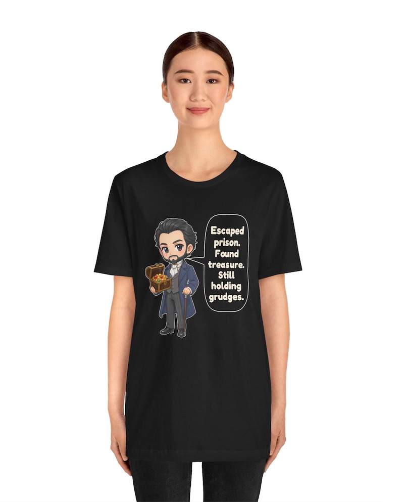 May include: A black t-shirt featuring a cartoon illustration of a bearded man in a blue coat holding a treasure chest. A speech bubble next to the character contains the text: Escaped prison. Found treasure. Still holding grudges.