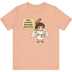 May include: A peach-colored t-shirt featuring a graphic of a cartoon angel with dark hair, white wings, and a crooked halo. The angel holds a scroll and waves, with a speech bubble containing the text: Halo crooked. Message delivered.