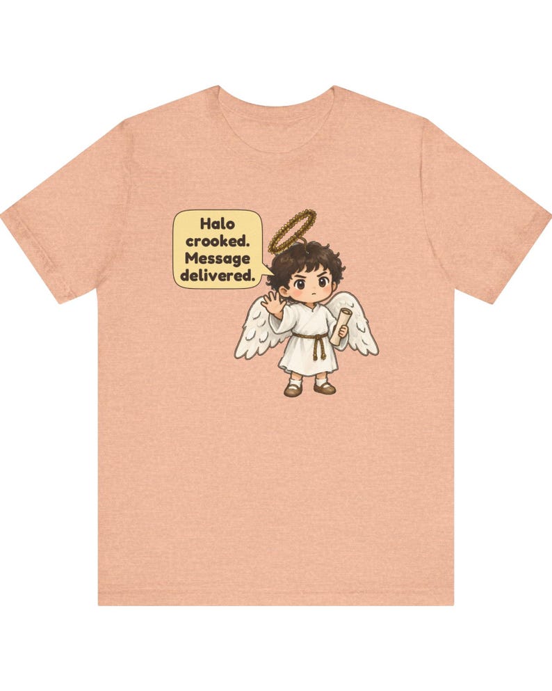 May include: A peach-colored t-shirt featuring a graphic of a cartoon angel with dark hair, white wings, and a crooked halo. The angel holds a scroll and waves, with a speech bubble containing the text: Halo crooked. Message delivered.