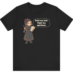 May include: A black t-shirt featuring a cartoon illustration of a person with dark hair, wearing a brown dress and black apron, holding a quill and paper. A speech bubble contains the text: Sold my hair. Kept my opinions.