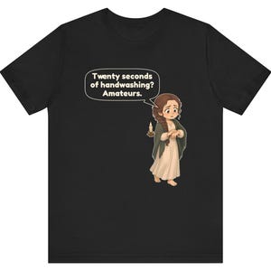 Op de afbeelding: Een zwart t-shirt met een cartoonillustratie van een persoon met lang bruin haar in een vlecht, gekleed in een cr&egrave;mekleurige jurk en een donkere sjaal, die een brandende kaars vasthoudt. Een tekstballon bevat de tekst: Twenty seconds of handwashing? Amateurs.