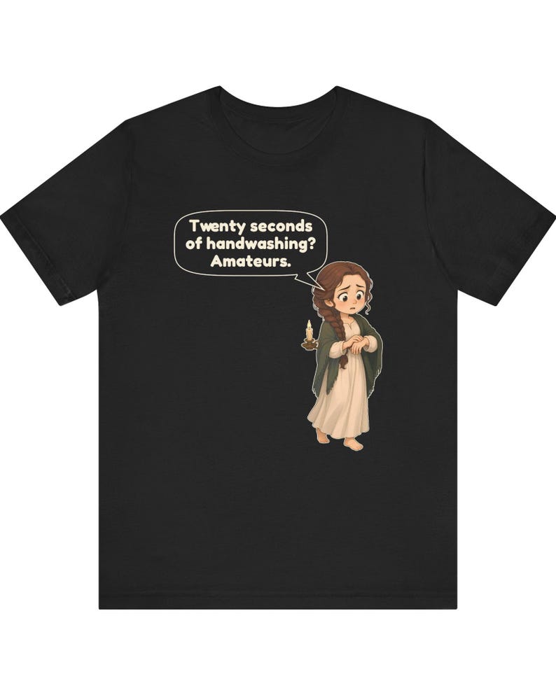 Op de afbeelding: Een zwart t-shirt met een cartoonillustratie van een persoon met lang bruin haar in een vlecht, gekleed in een cr&egrave;mekleurige jurk en een donkere sjaal, die een brandende kaars vasthoudt. Een tekstballon bevat de tekst: Twenty seconds of handwashing? Amateurs.