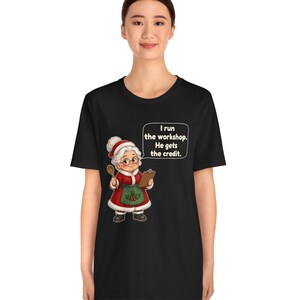K&ouml;nnte beinhalten: Ein schwarzes Kurzarm-T-Shirt mit einer Grafik einer &auml;lteren Frau in einem rot-wei&szlig;en Kleid mit gr&uuml;ner Sch&uuml;rze, die einen Holzl&ouml;ffel und ein Klemmbrett h&auml;lt. Eine Sprechblase enth&auml;lt den Text: I run the workshop. He gets the credit.