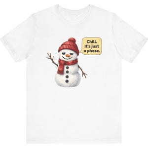 Pode incluir: Uma t-shirt branca com um gr&aacute;fico de um boneco de neve a usar um gorro e um cachecol de malha vermelhos. O boneco de neve tem bra&ccedil;os de pau, um nariz de cenoura e bot&otilde;es pretos. Um bal&atilde;o de fala amarelo cont&eacute;m o texto, Chill. It's just a phase.