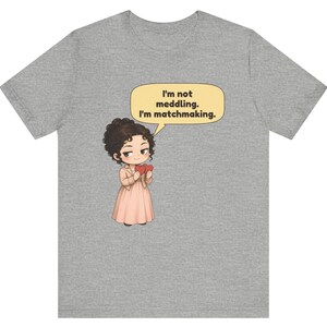 K&ouml;nnte beinhalten: Ein graumeliertes T-Shirt mit einer Cartoon-Illustration einer Person mit dunklen lockigen Haaren, die ein hellrosa Kleid und eine Jacke tr&auml;gt und zwei rote Herzen h&auml;lt, mit einer Sprechblase, die besagt, I'm not meddling. I'm matchmaking.