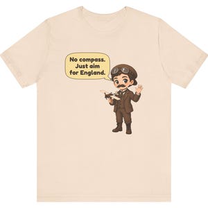 May include: A light beige t-shirt featuring a cartoon illustration of a pilot with a mustache, wearing a brown suit, cap, and goggles. The pilot holds a small airplane model and a speech bubble with the text: No compass. Just aim for England.