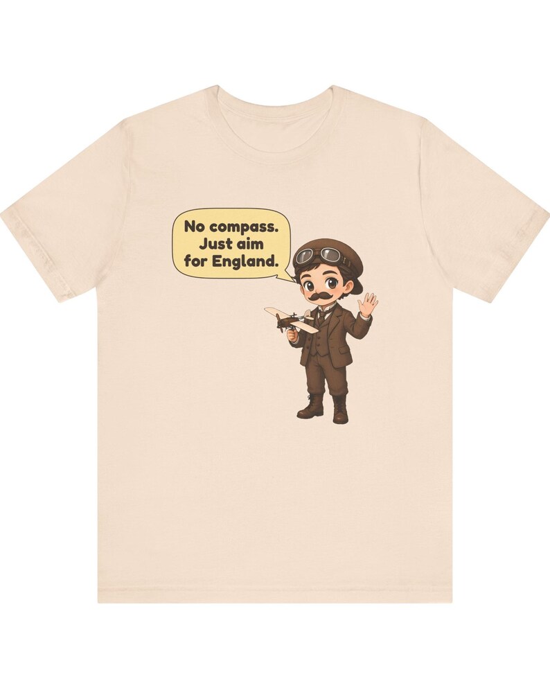 May include: A light beige t-shirt featuring a cartoon illustration of a pilot with a mustache, wearing a brown suit, cap, and goggles. The pilot holds a small airplane model and a speech bubble with the text: No compass. Just aim for England.