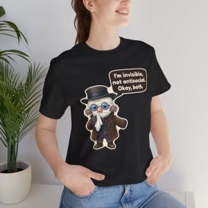 May include: A black short-sleeve t-shirt featuring a cartoon illustration of a bandaged figure wearing a top hat, glasses, and a brown coat, holding a white cloth. A speech bubble contains the text: I'm invisible, not antisocial. Okay, both.