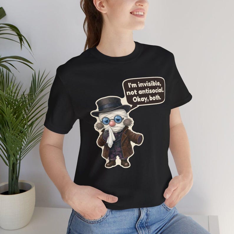 May include: A black short-sleeve t-shirt featuring a cartoon illustration of a bandaged figure wearing a top hat, glasses, and a brown coat, holding a white cloth. A speech bubble contains the text: I'm invisible, not antisocial. Okay, both.