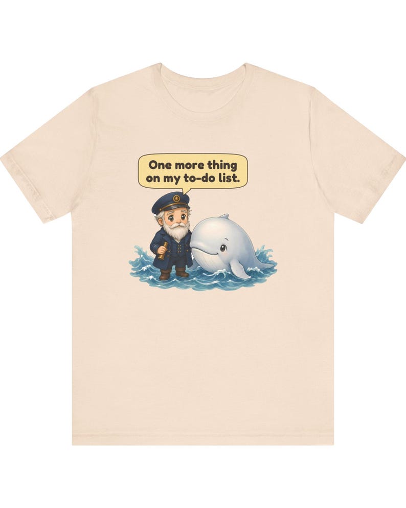 May include: A light beige t-shirt featuring a graphic of a bearded sea captain in a navy uniform standing next to a white whale in blue ocean waves. Above the characters, a speech bubble contains the text, One more thing on my to-do list.