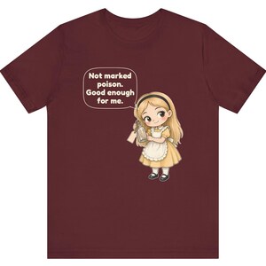 May include: A maroon t-shirt featuring a cartoon illustration of a blonde character in a yellow dress and white apron holding a glass bottle. A speech bubble above the character contains the text: Not marked poison. Good enough for me.