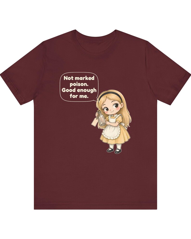 May include: A maroon t-shirt featuring a cartoon illustration of a blonde character in a yellow dress and white apron holding a glass bottle. A speech bubble above the character contains the text: Not marked poison. Good enough for me.
