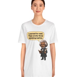 Puede incluir: Una camiseta blanca con una ilustraci&oacute;n de dibujos animados de Frederick Douglass sosteniendo un libro y una pluma. Un globo de texto sobre la figura contiene el texto: Learned to read. Then wrote three autobiographies.