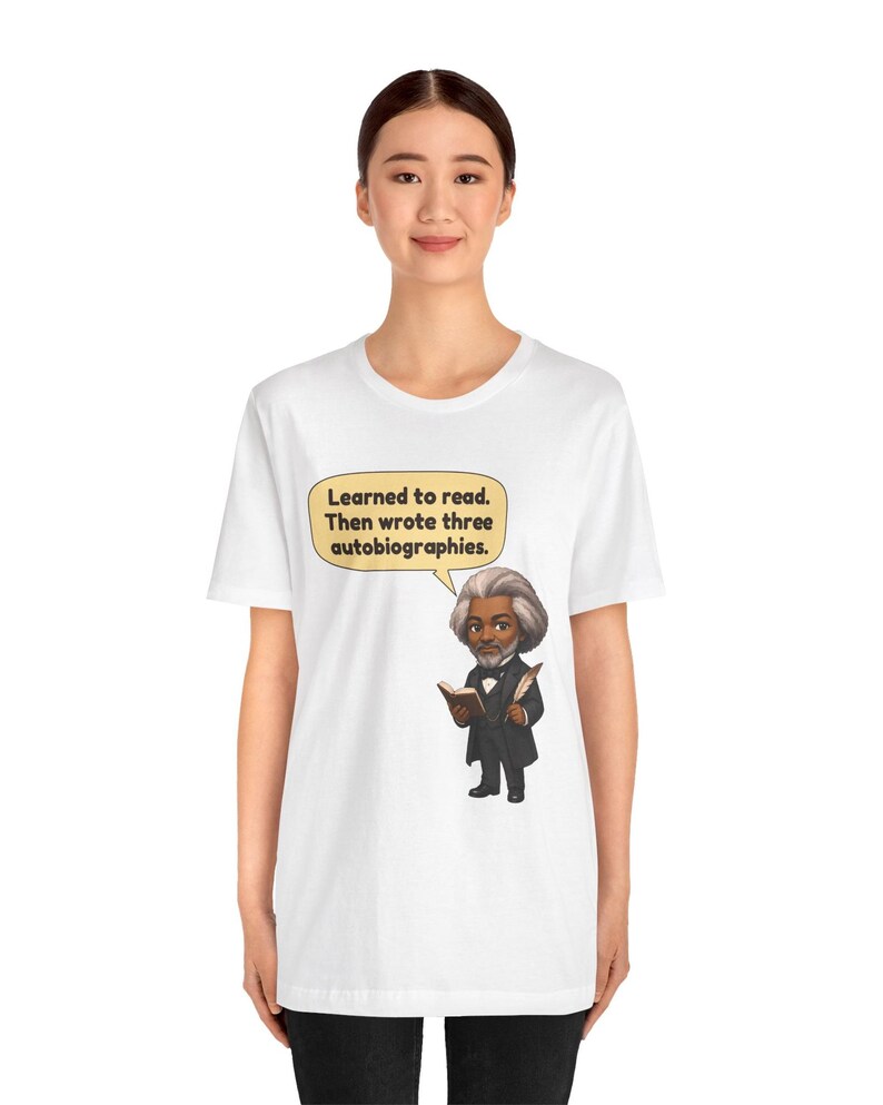 Puede incluir: Una camiseta blanca con una ilustraci&oacute;n de dibujos animados de Frederick Douglass sosteniendo un libro y una pluma. Un globo de texto sobre la figura contiene el texto: Learned to read. Then wrote three autobiographies.