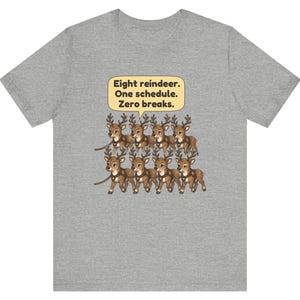 K&ouml;nnte beinhalten: Ein graues, meliertes T-Shirt mit einer Grafik von acht braunen Rentieren in zwei Reihen. &Uuml;ber den Rentieren befindet sich eine gelbe Sprechblase mit dem schwarzen Text: Eight reindeer. One schedule. Zero breaks.