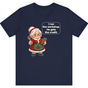 K&ouml;nnte beinhalten: Ein marineblaues T-Shirt mit einer Cartoon-Illustration einer &auml;lteren Frau in einem rot-wei&szlig;en Outfit im Weihnachtsmann-Stil mit gr&uuml;ner Sch&uuml;rze, die einen Holzl&ouml;ffel und ein Klemmbrett h&auml;lt, mit einer Sprechblase, die sagt, I run the workshop. He gets the credit.
