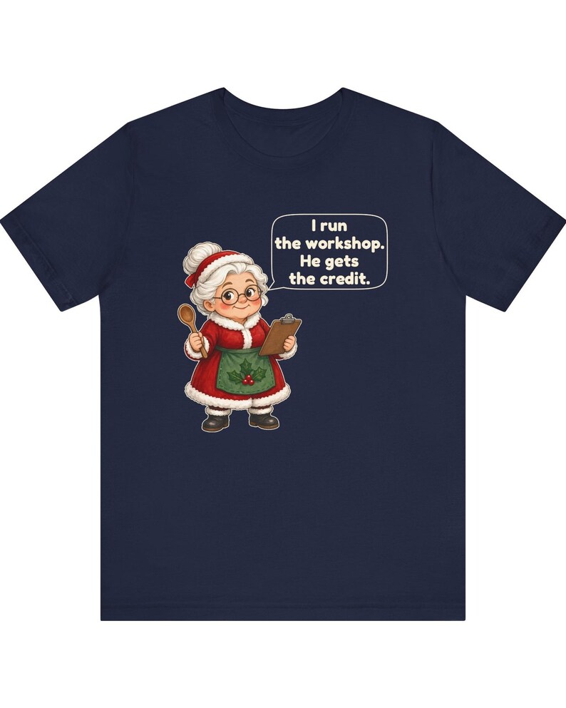 K&ouml;nnte beinhalten: Ein marineblaues T-Shirt mit einer Cartoon-Illustration einer &auml;lteren Frau in einem rot-wei&szlig;en Outfit im Weihnachtsmann-Stil mit gr&uuml;ner Sch&uuml;rze, die einen Holzl&ouml;ffel und ein Klemmbrett h&auml;lt, mit einer Sprechblase, die sagt, I run the workshop. He gets the credit.
