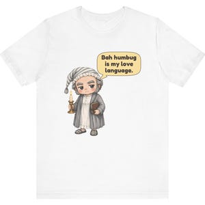 Puede incluir: Una camiseta blanca con una ilustraci&oacute;n de dibujos animados de un personaje con gorro de dormir y bata que sostiene una vela y un libro. Un bocadillo junto al personaje contiene el texto: Bah humbug is my love language.