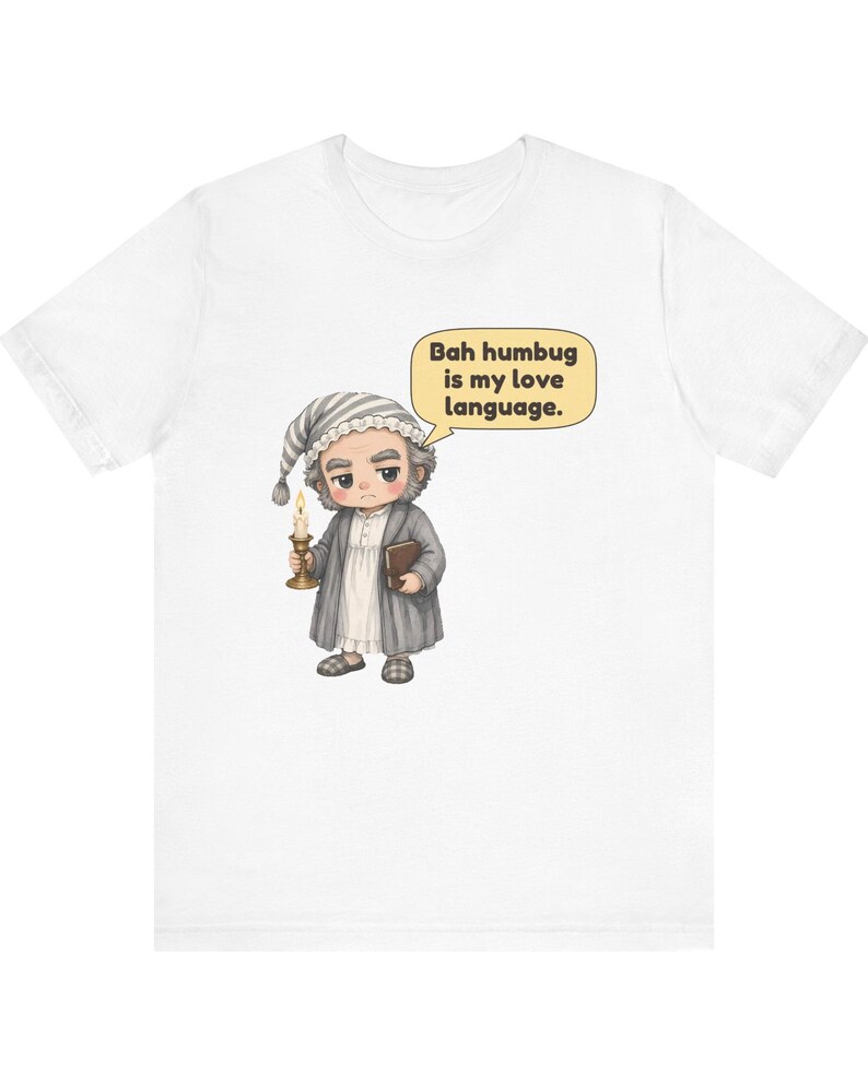 Puede incluir: Una camiseta blanca con una ilustraci&oacute;n de dibujos animados de un personaje con gorro de dormir y bata que sostiene una vela y un libro. Un bocadillo junto al personaje contiene el texto: Bah humbug is my love language.
