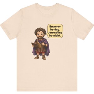 K&ouml;nnte beinhalten: Ein hellbeiges T-Shirt mit einer Cartoon-Illustration eines b&auml;rtigen Kaisers in r&ouml;mischer R&uuml;stung und violettem Umhang, der ein Tagebuch und einen Stift h&auml;lt. Eine Sprechblase neben der Figur enth&auml;lt den Text: Emperor by day. Journaling by night.