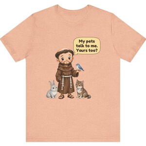 May include: A peach-colored t-shirt featuring a cartoon illustration of a monk in a brown robe standing with a gray rabbit, a small wolf, and a blue bird. A speech bubble above the monk contains the text, My pets talk to me. Yours too?
