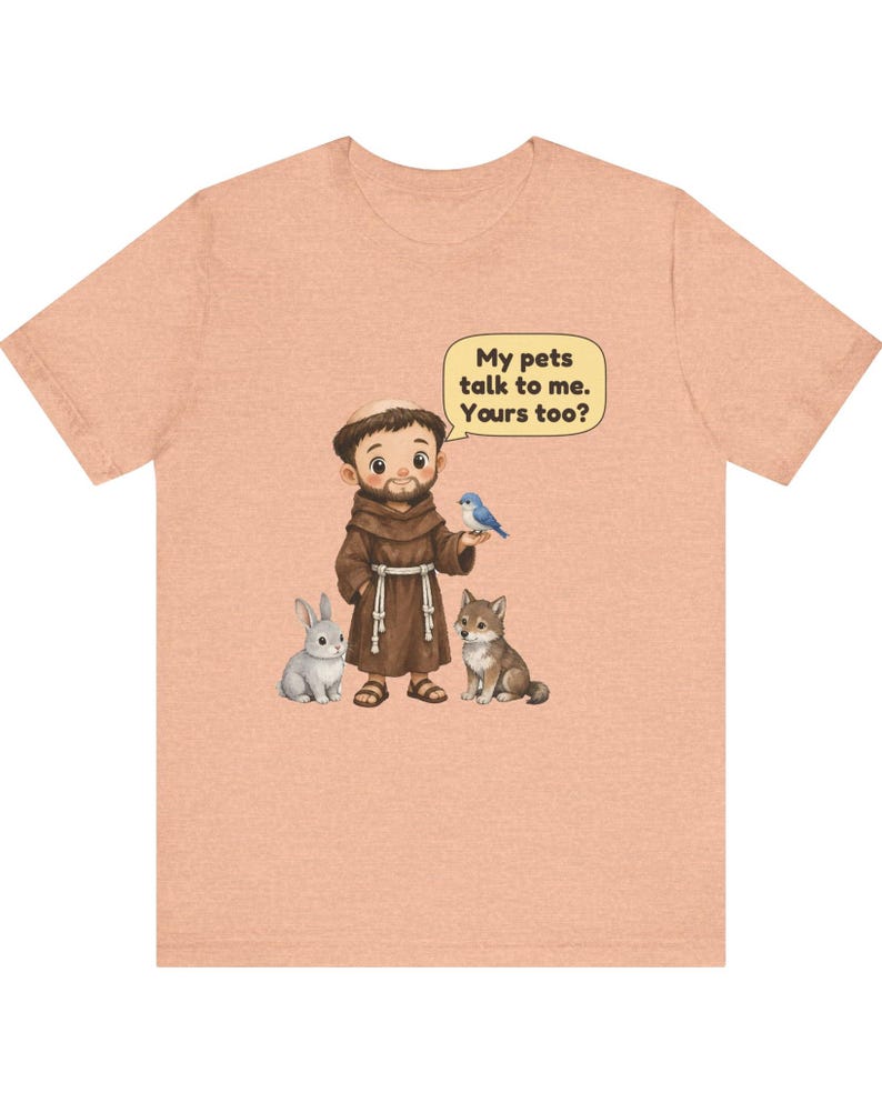 May include: A peach-colored t-shirt featuring a cartoon illustration of a monk in a brown robe standing with a gray rabbit, a small wolf, and a blue bird. A speech bubble above the monk contains the text, My pets talk to me. Yours too?