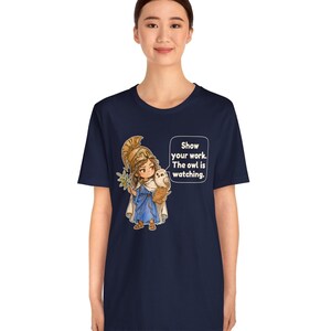 Puede incluir: Una camiseta azul marino con un gr&aacute;fico de un personaje de dibujos animados con casco dorado y t&uacute;nica azul, sosteniendo una rama de olivo y un b&uacute;ho. Un bocadillo contiene el texto: Show your work. The owl is watching.