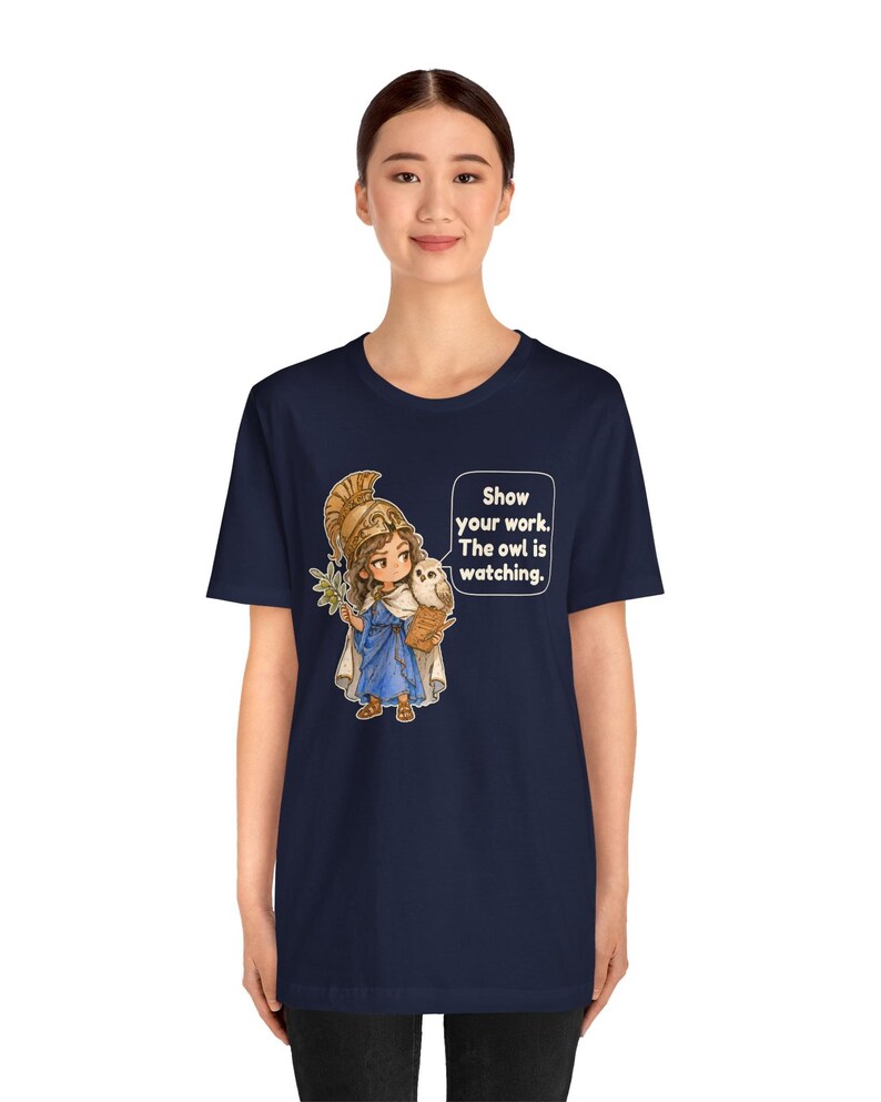 Puede incluir: Una camiseta azul marino con un gr&aacute;fico de un personaje de dibujos animados con casco dorado y t&uacute;nica azul, sosteniendo una rama de olivo y un b&uacute;ho. Un bocadillo contiene el texto: Show your work. The owl is watching.