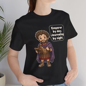 K&ouml;nnte beinhalten: Ein schwarzes T-Shirt mit einer Cartoon-Illustration eines b&auml;rtigen Mannes in r&ouml;mischer R&uuml;stung und violettem Umhang, der einen Stift und ein Tagebuch h&auml;lt. Eine Sprechblase enth&auml;lt den Text: Emperor by day. Journaling by night.