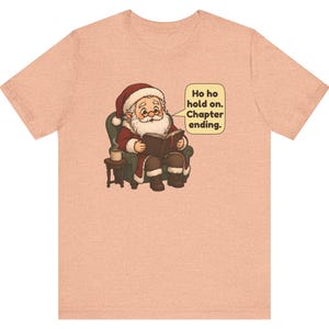 Op de afbeelding: Een perzikkleurig t-shirt met een afbeelding van de Kerstman die in een groene fauteuil zit, een boek leest en een kopje koffie op een bijzettafeltje heeft staan. Een tekstballon bevat de tekst: Ho ho hold on. Chapter ending.