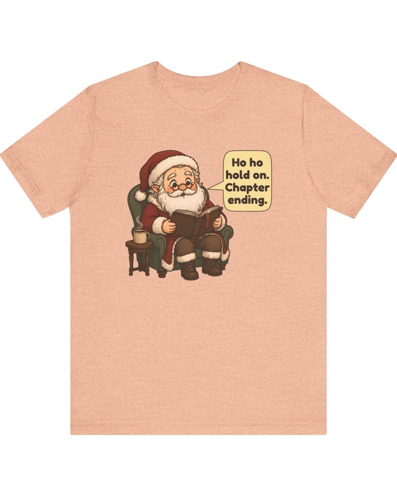 Op de afbeelding: Een perzikkleurig t-shirt met een afbeelding van de Kerstman die in een groene fauteuil zit, een boek leest en een kopje koffie op een bijzettafeltje heeft staan. Een tekstballon bevat de tekst: Ho ho hold on. Chapter ending.