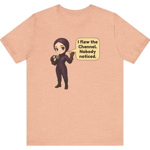 Puede incluir: Una camiseta color melocot&oacute;n con un personaje de dibujos animados con traje de vuelo morado y gafas. Un bocadillo junto al personaje contiene el texto: I flew the Channel. Nobody noticed. El dise&ntilde;o est&aacute; centrado en la parte delantera.
