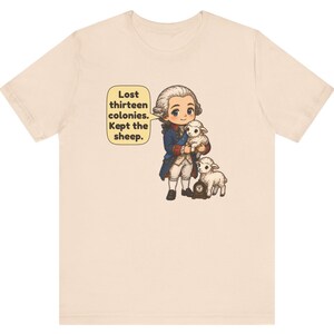Peut inclure: Un t-shirt beige clair avec une illustration de style dessin anim&eacute; repr&eacute;sentant un personnage historique en manteau bleu et perruque blanche tenant un agneau, avec un autre agneau et une petite horloge. Une bulle contient le texte : Lost thirteen colonies. Kept the sheep.