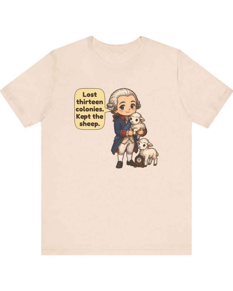 Peut inclure: Un t-shirt beige clair avec une illustration de style dessin anim&eacute; repr&eacute;sentant un personnage historique en manteau bleu et perruque blanche tenant un agneau, avec un autre agneau et une petite horloge. Une bulle contient le texte : Lost thirteen colonies. Kept the sheep.