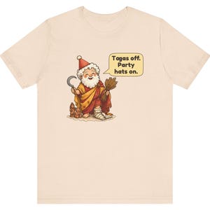 May include: A light beige t-shirt featuring a cartoon illustration of a bearded figure wearing a red and yellow robe, a red hat, and holding a sickle and wheat. A speech bubble contains the text: Togas off. Party hats on.